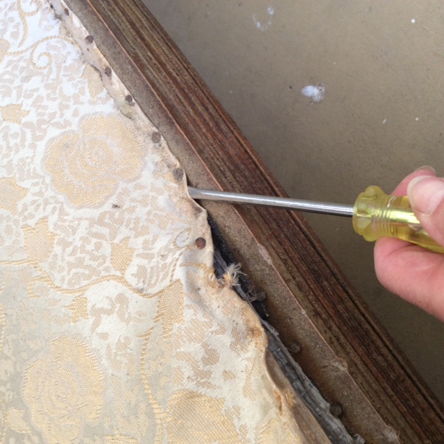 Close up image of a hand using a flathead screwdriver to remove the old cushion nails from the frame of the loveseat.