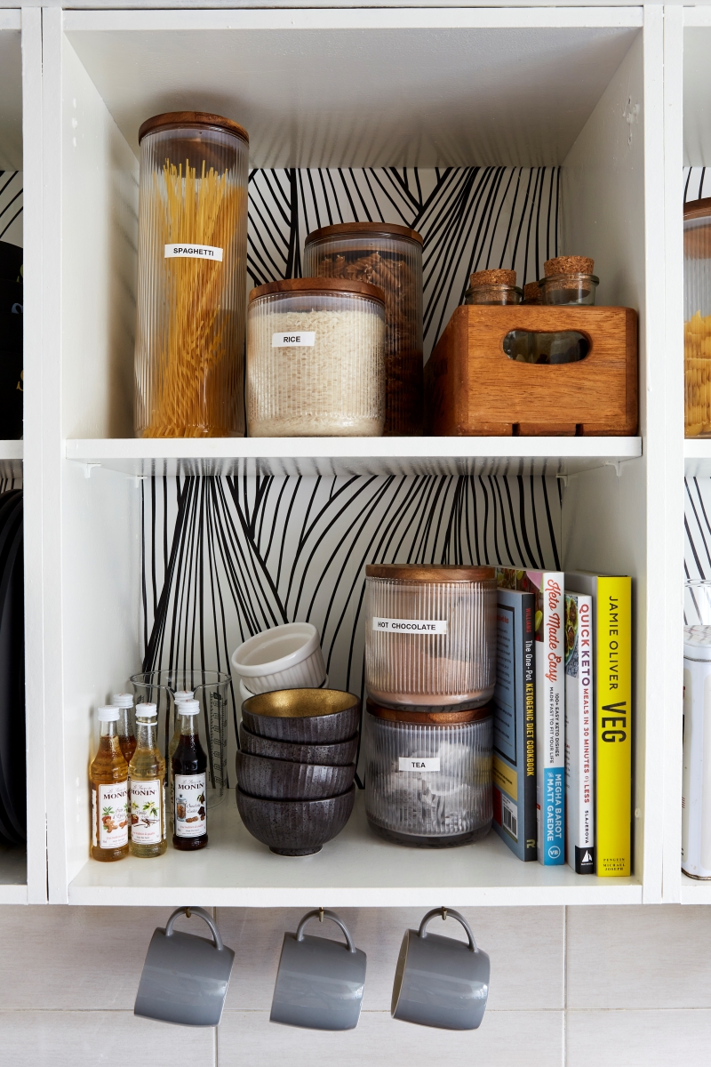 close up image of cabinet in the renter friendly kitchen painted white with without the door and graphic black and white wallpaper on the back and pantry items and cookbooks on the shelves