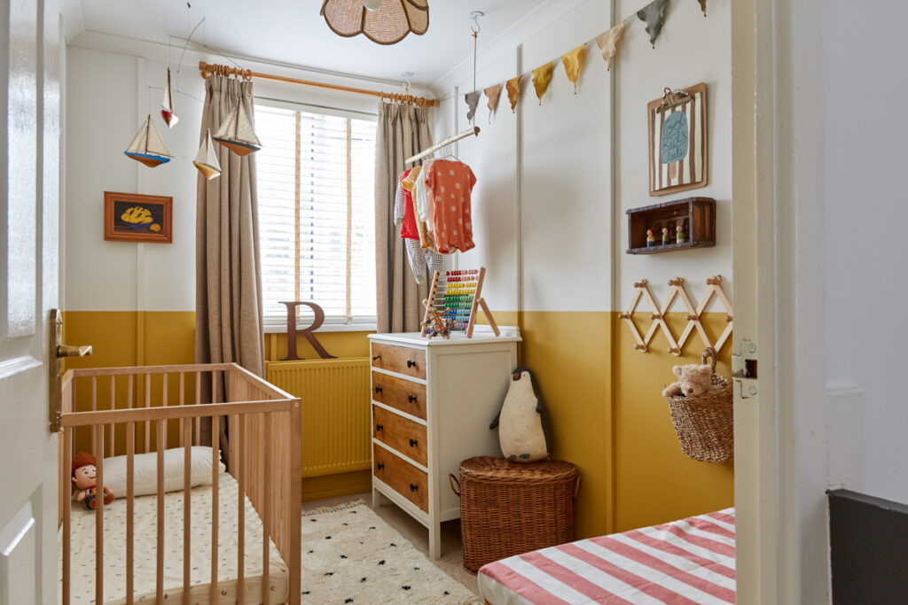 small nursery reveal showing a half painted wall in a warm yellow. there is a cot on the left and a chest of drawers on the left, a hanging rail above this made from a branch. Some cute furnishings can be see too.