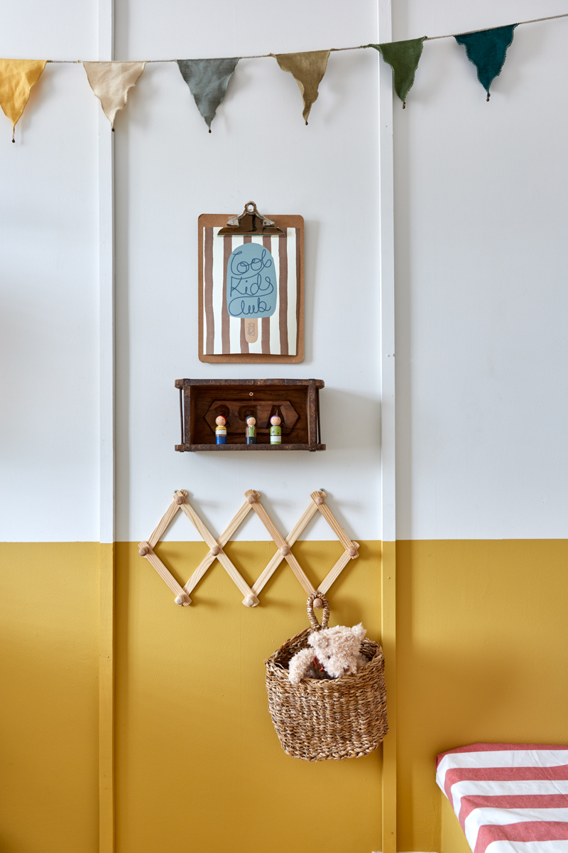 the panelling is shown clearly here, bunting at the top of the image with flags in earthy tones, a wooden box as a shelf, a clipboard to hang artwork, a small bear in a basket hung on a diamond wooden rail
