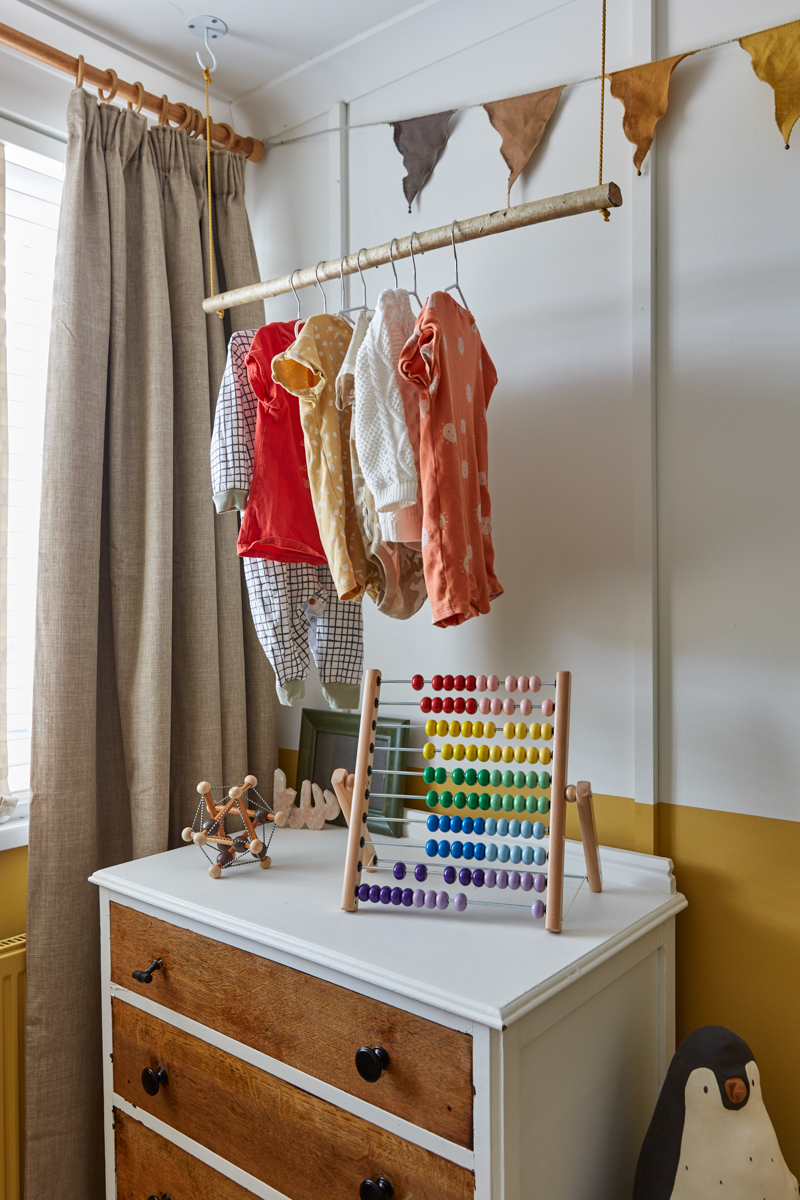 A wooden branch pole is suspended by string to create a small hanging rail. Taupe coloured natural thick cotton curtains hang in the window, bunting above the rail on the wall and below it a chest of drawers sit with cute wooden toys