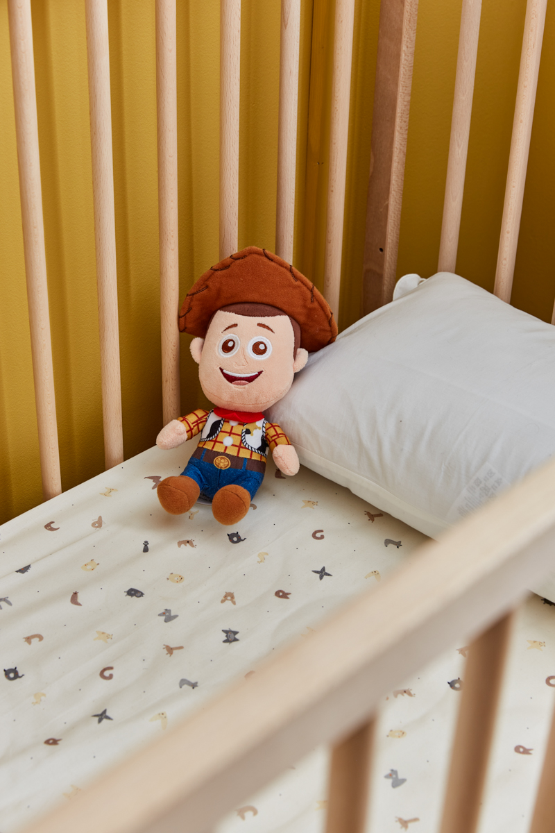 the yellow wall can be seen through the cot rails. on the bedding which is a small alphabet print lays a cute Woody from Toy Story soft toy, matching the decor perfectly 