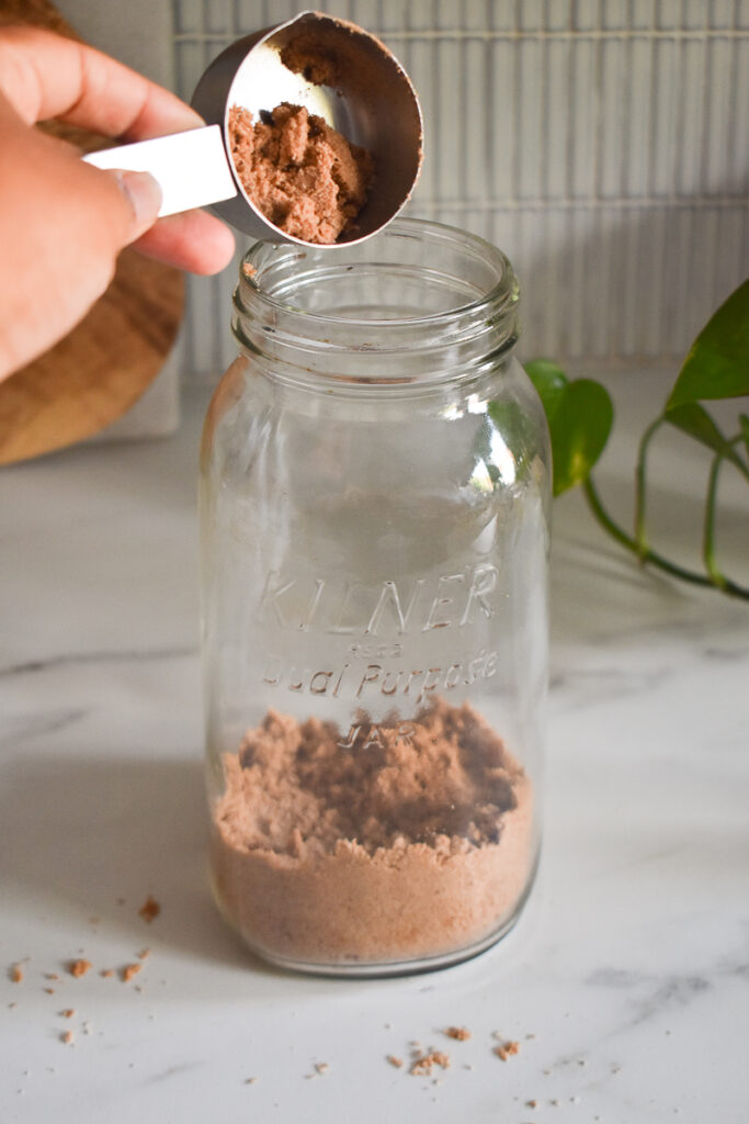 a mason jar on a marble type white counter and brown hand scooping in sand with a silver measuring spoon