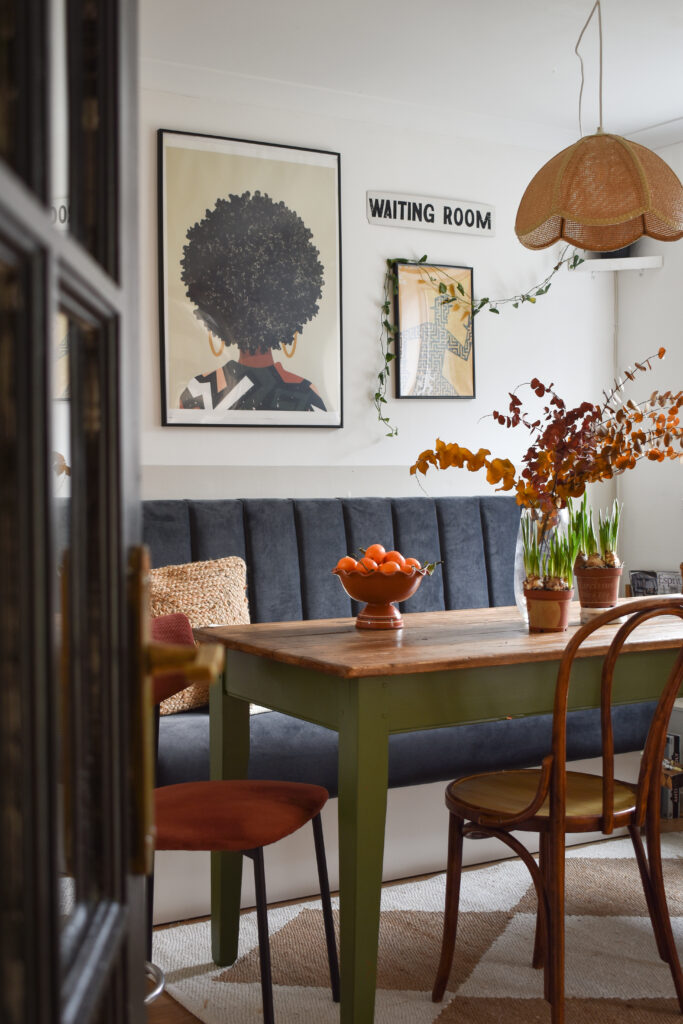a vintage modern dining room with warm tones. A painting of a Black woman is hung on the wall above the banquet seating. Her afro is perfectly rounded. A rattan vintage lampshade hangs above a vintage table with a sanded top and dark green legs. 