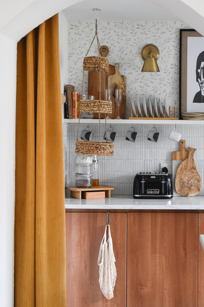 a peek into the kitchen through mustard yellow thick ribbed curtain. Open shelving with plates and cutting boards sit above a faux marble worktop. There is a black toaster and hot chocolate station on top and mugs hung from the shelving. 